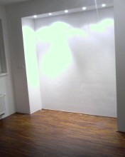 Pusta wnęka w ścianie, oświetlona od góry punktowymi lampami, z ciemną, błyszczącą podłogą. Widoczny fragment okna i grzejnika. Ściany świeżo pomalowane na biało.